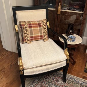 Puede incluir: Una silla negra antigua con detalles dorados y un asiento color crema. Un coj&iacute;n a cuadros con flecos descansa sobre el asiento. La silla est&aacute; sobre una alfombra estampada, con un suelo de madera y un armario al fondo.