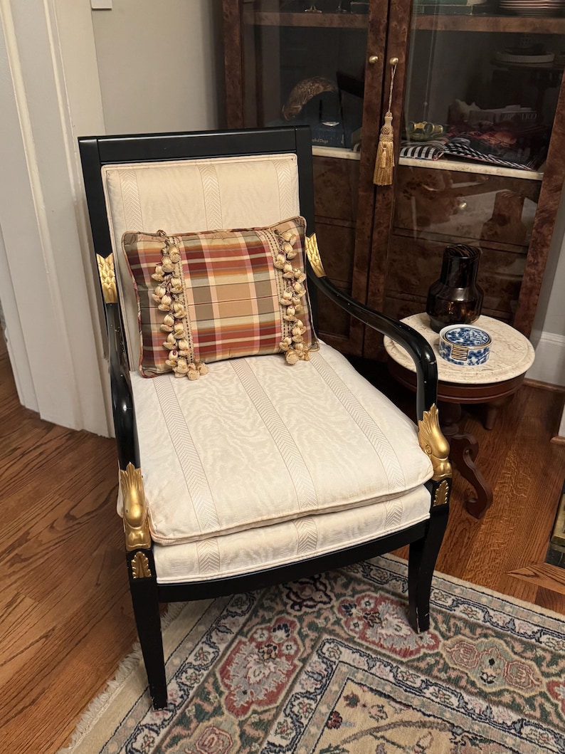 Puede incluir: Una silla negra antigua con detalles dorados y un asiento color crema. Un coj&iacute;n a cuadros con flecos descansa sobre el asiento. La silla est&aacute; sobre una alfombra estampada, con un suelo de madera y un armario al fondo.