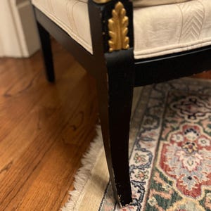 Puede incluir: Primer plano de una pata de silla negra con detalles de pan de oro. La silla tiene un coj&iacute;n color crema con un dise&ntilde;o estampado. La silla est&aacute; sobre una alfombra estampada y un suelo de madera.