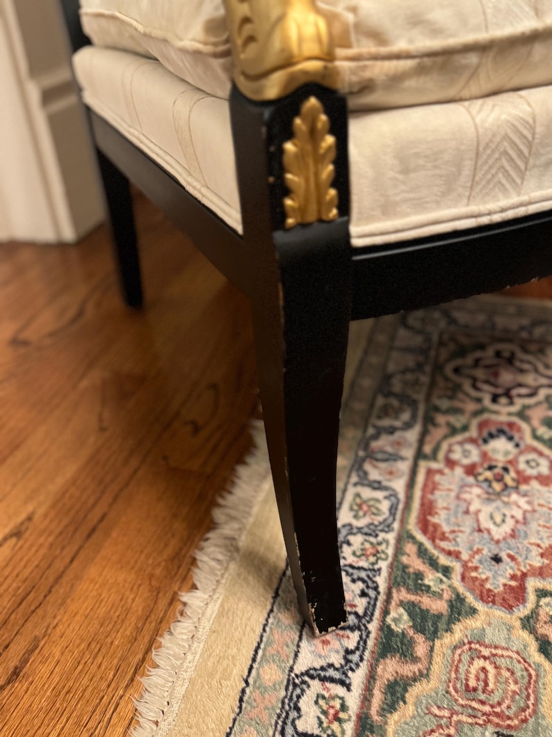 Puede incluir: Primer plano de una pata de silla negra con detalles de pan de oro. La silla tiene un coj&iacute;n color crema con un dise&ntilde;o estampado. La silla est&aacute; sobre una alfombra estampada y un suelo de madera.