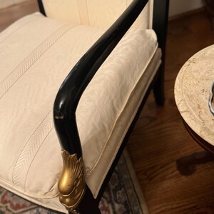 Puede incluir: Un elegante sill&oacute;n negro con detalles dorados. El reposabrazos curvo presenta un elemento decorativo dorado. El asiento y el respaldo est&aacute;n tapizados en tela color crema con un sutil dise&ntilde;o de rayas. La silla forma parte de un conjunto de muebles.
