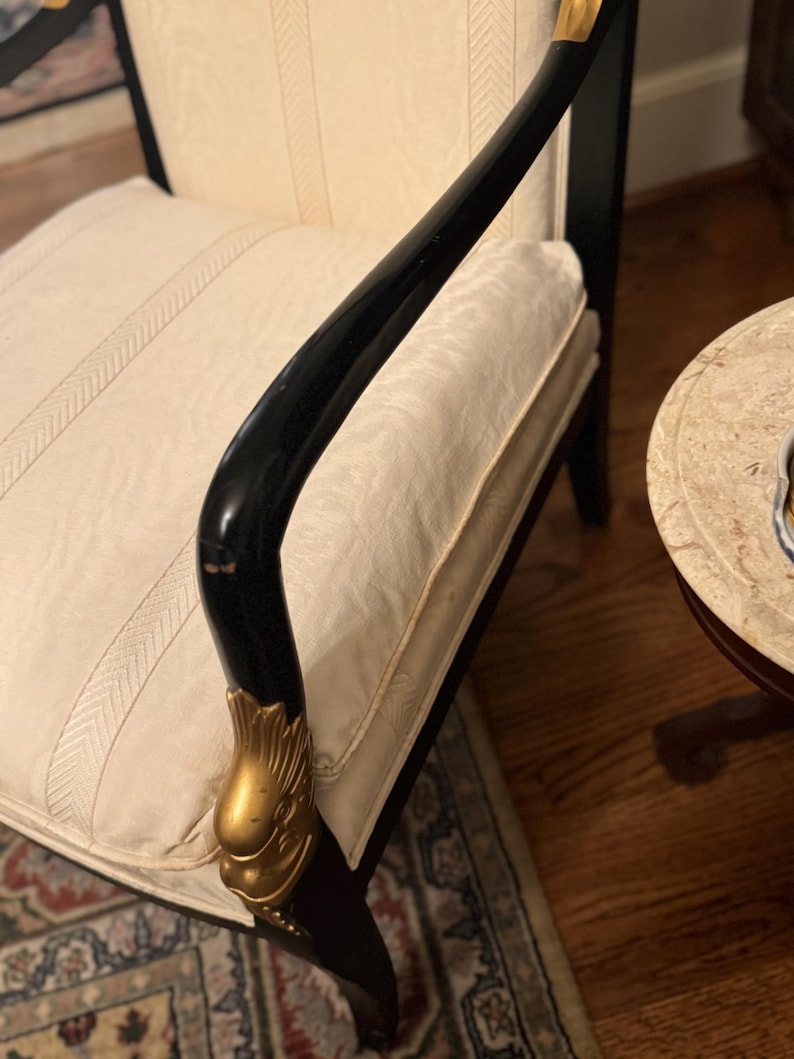 Puede incluir: Un elegante sill&oacute;n negro con detalles dorados. El reposabrazos curvo presenta un elemento decorativo dorado. El asiento y el respaldo est&aacute;n tapizados en tela color crema con un sutil dise&ntilde;o de rayas. La silla forma parte de un conjunto de muebles.