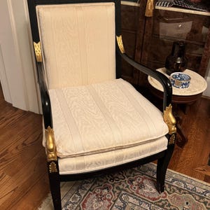 Puede incluir: Un sill&oacute;n antiguo con estructura negra y tapicer&iacute;a crema. La silla presenta detalles decorativos dorados en los brazos y las patas. El asiento y el respaldo tienen un patr&oacute;n de rayas verticales.
