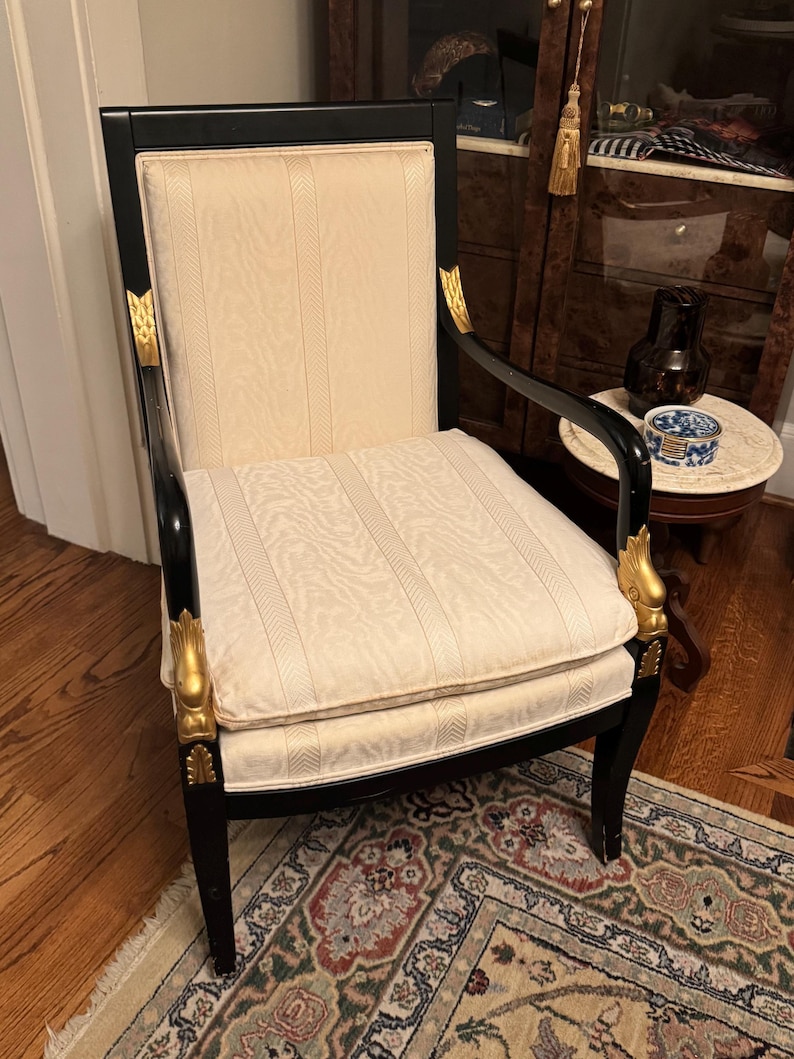 Puede incluir: Un sill&oacute;n antiguo con estructura negra y tapicer&iacute;a crema. La silla presenta detalles decorativos dorados en los brazos y las patas. El asiento y el respaldo tienen un patr&oacute;n de rayas verticales.