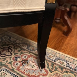 Puede incluir: Primer plano de una pata de silla negra con detalles dorados. La silla tiene un asiento color crema con un dise&ntilde;o estampado. La pata se apoya en una alfombra estampada con tonos rojos, verdes y beige.