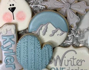 May include: A collection of winter-themed sugar cookies on a white tray. The cookies include a polar bear, a snowflake, a mitten, a mountain scene, and a boot with the name "Kyler". The cookies are decorated with white, light blue, and silver icing.