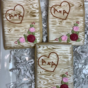 May include: Three decorated sugar cookies with a woodgrain design. Each cookie features a heart with the initials "A*A" and a floral design in the corner. The cookies are on a white plate with white shredded paper.
