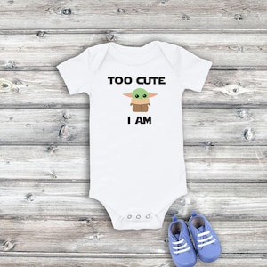 Op de afbeelding: Witte baby romper met een groen alien-figuur en de tekst "TOO CUTE I AM".