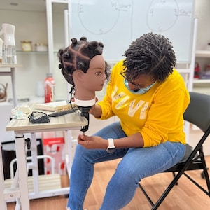 Puede incluir: Una persona con el pelo castaño oscuro en trenzas se sienta en una silla y trabaja en una cabeza de maniquí con un peinado de trenzas. La cabeza de maniquí está unida a un soporte. La persona lleva una camiseta amarilla con un gráfico blanco, pantalones vaqueros azules y zapatillas blancas.