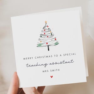 May include: A white Christmas card featuring a hand-drawn green Christmas tree with red and gold baubles. The text reads "Merry Christmas to a special teaching assistant" and "Mrs Smith".