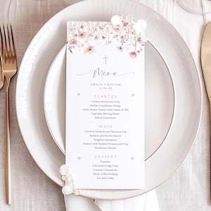 Puede incluir: Una tarjeta de menú para el bautizo de Amelia, con un diseño floral en rosa y blanco. El menú incluye entrantes, platos principales y postres, como sopa cremosa de champiñones y pastel de fudge de chocolate. La tarjeta está sobre un plato blanco con cubiertos dorados.