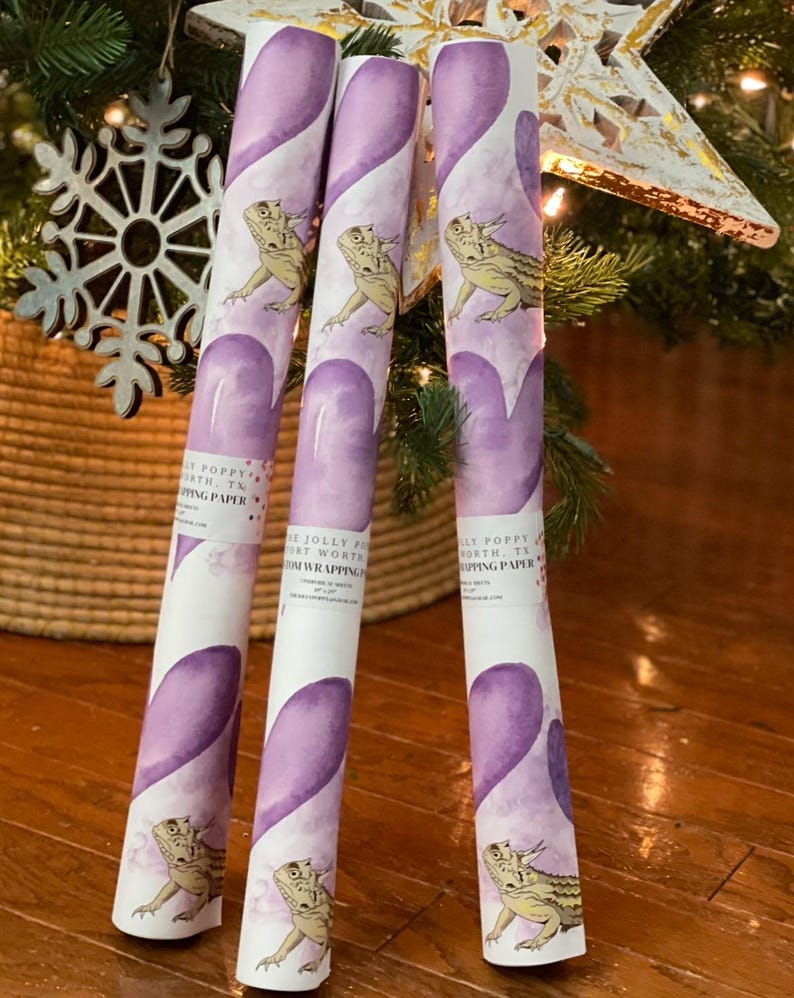 May include: Three rolls of purple and white wrapping paper with a watercolor design of hearts and a lizard. The paper is labeled "The Jolly Poppy Fort Worth, TX Gift Wrapping".