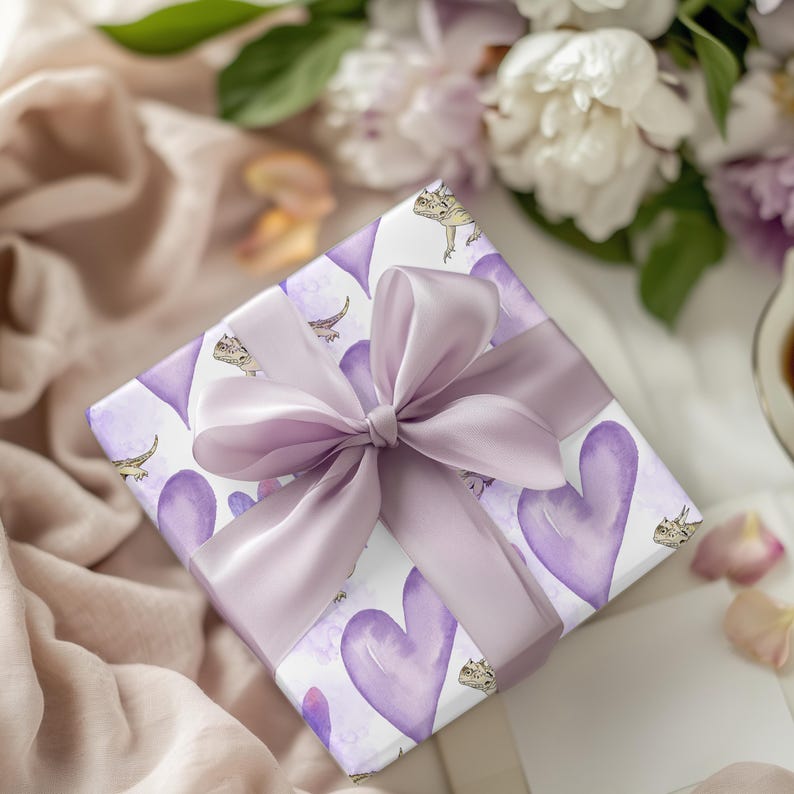May include: A white gift box with a purple and white patterned design featuring hearts and small lizards. The box is tied with a large purple satin bow.