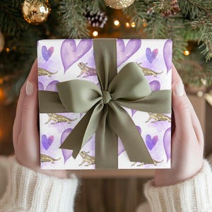 May include: A gift wrapped in white paper with a green bow. The paper has a repeating pattern of purple hearts and brown bearded dragons.