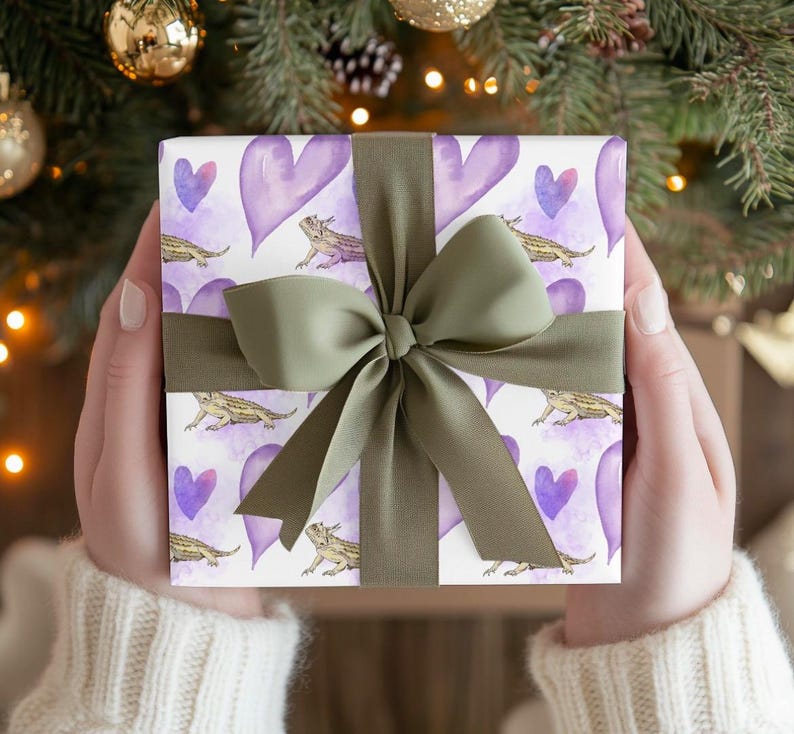 May include: A gift wrapped in white paper with a green bow. The paper has a repeating pattern of purple hearts and brown bearded dragons.