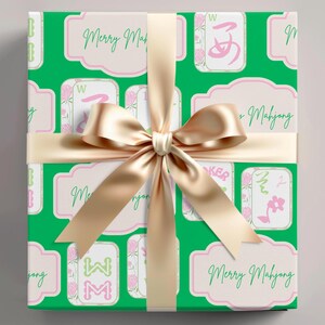 以下が含まれることがあります： 麻雀牌のパターンと「Merry Mahjong」の文字が入った緑色の包装紙で包まれたギフト。 金色のサテンリボンとリボンがパッケージを飾っています。 ギフト包装は、ピンクのアクセントが付いた白い背景です。