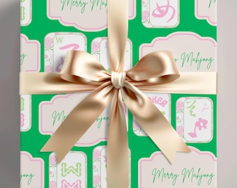 May include: A wrapped gift with green wrapping paper featuring a pattern of Mahjong tiles and the words "Merry Mahjong." A gold satin ribbon and bow adorn the package. The gift wrap has a white background with pink accents.