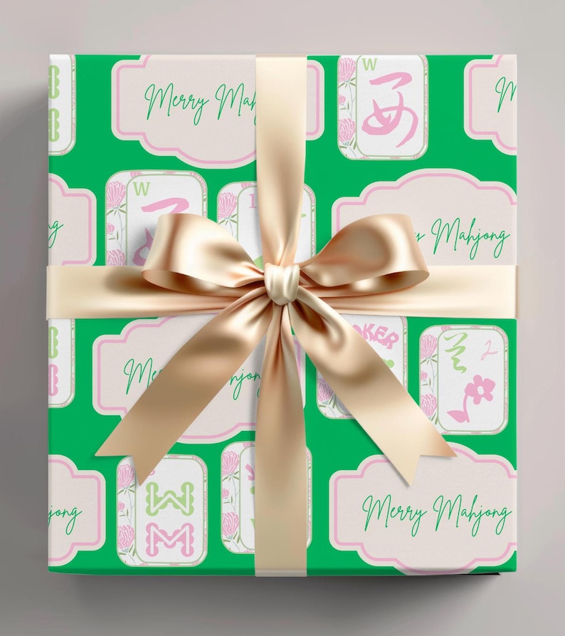 以下が含まれることがあります： 麻雀牌のパターンと「Merry Mahjong」の文字が入った緑色の包装紙で包まれたギフト。 金色のサテンリボンとリボンがパッケージを飾っています。 ギフト包装は、ピンクのアクセントが付いた白い背景です。