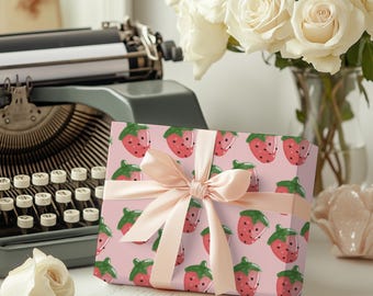 May include: A pink gift box with a pink ribbon and a strawberry pattern. The box is sitting on a table with a typewriter and white roses in the background.