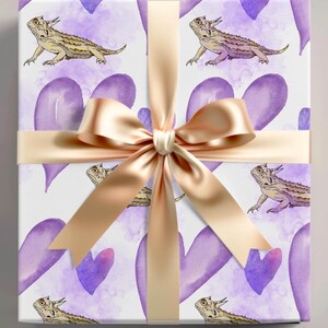 May include: Gift wrap featuring a repeating pattern of purple watercolor hearts and cartoon horned lizards. The paper is tied with a gold satin ribbon.