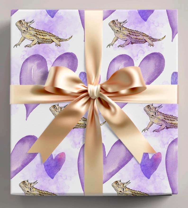 May include: Gift wrap featuring a repeating pattern of purple watercolor hearts and cartoon horned lizards. The paper is tied with a gold satin ribbon.
