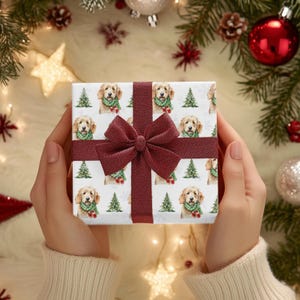以下が含まれることがあります： 白い背景に、緑のスカーフとクリスマスツリーを身に着けたゴールデンレトリバーの繰り返し模様が特徴の、包まれたギフト。ギフトはバーガンディのリボンとリボンで結ばれています。背景にはクリスマスの飾りがあります。