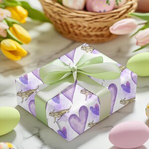 May include: A white gift box with a green ribbon and bow. The box is wrapped in paper with a pattern of purple hearts and brown lizards.