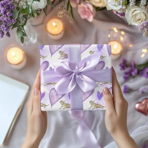 May include: A purple gift box with a white background and a purple ribbon. The box is decorated with a pattern of purple hearts and small lizards.