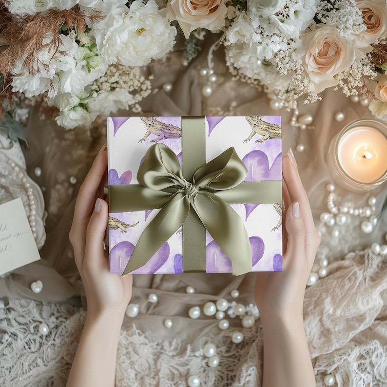 May include: A gift box wrapped in white paper with a purple and white heart pattern and a green bow. The box features a watercolor illustration of a bearded dragon.