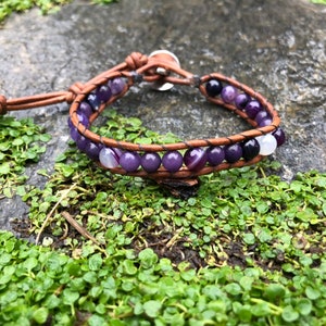 May include: A brown leather bracelet with purple and white agate beads. The bracelet has a silver clasp and is adjustable.