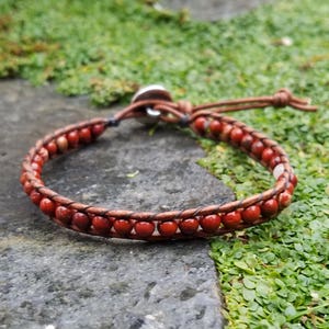 Puede incluir: Un brazalete de cuero marrón con cuentas de piedras preciosas rojas. El brazalete tiene un cierre plateado y es ajustable.