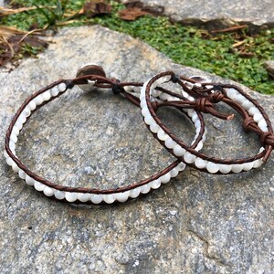 May include: Two leather bracelets with white shell beads. The bracelets are brown leather with white beads and are tied together with a knot.