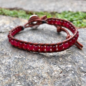 以下が含まれることがあります： 赤い宝石のビーズが付いた茶色の革のラップブレスレット。ブレスレットには銀色の留め具が付いています。