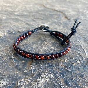 May include: A wrap bracelet featuring small, round, reddish-brown beads. The beads are strung on a black leather band. The bracelet has a silver-colored button clasp and a knotted leather closure. The bracelet is designed to wrap around the wrist.