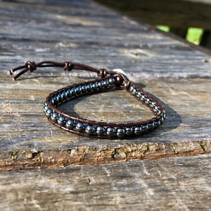 May include: A wrap bracelet featuring dark gray beads and brown leather. The bracelet is made with two rows of beads and a braided leather band. The bracelet is tied with a leather cord.