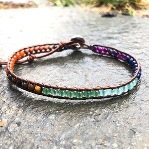 May include: A wrap bracelet with a brown leather band and colorful beads. The beads transition from orange to green to blue and purple. The bracelet is secured with a silver button clasp.