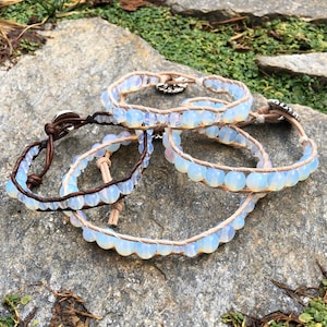 May include: Four leather wrap bracelets with white opal beads. The bracelets are stacked on top of each other on a gray rock.