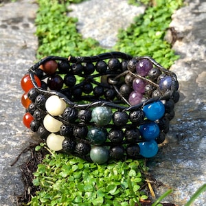May include: A multi-layered bracelet with a variety of colourful beads. The bracelet incorporates black lava rock beads and a spectrum of gemstones, including red, white, green, blue, and purple. The bracelet is held together with black leather.