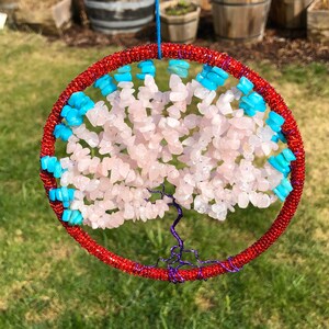 May include: A red beaded hoop with a wire tree of life design. The tree is decorated with pink and blue stones.