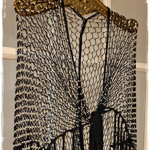 May include: Black crocheted shawl with fringe, adorned with small, light-colored beads. The shawl is displayed on a decorative gold-colored rod. The intricate net-like design and long fringe create a vintage, elegant look.