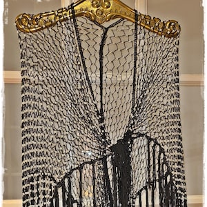 May include: A black, open-weave shawl with a decorative fringe. The shawl is adorned with small, off-white beads and hangs from a gold-colored, ornate hanger. The design features a net-like pattern.
