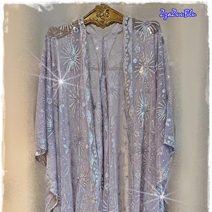 May include: A sheer, light purple open-front kimono-style jacket with silver sequin embellishments. The jacket features a floral and starburst pattern, with a decorative trim along the edges. The garment is hanging on a gold-colored hanger.