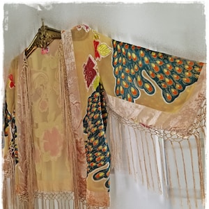 May include: A tan kimono-style jacket with a floral and peacock feather design. The jacket has long sleeves and is trimmed with fringe along the bottom and sleeves. The jacket is hanging on a gold hanger.