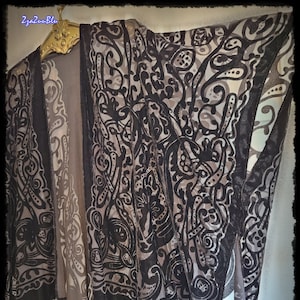 May include: A sheer, open-front kimono in shades of black, gray, and taupe, adorned with an intricate, swirling pattern. The kimono features a dark border and fringe along the hem. The garment is hanging on a gold-colored hanger.