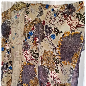 Op de afbeelding: Een beige kimono met bloemen en een donkerpaarse rand. De kimono heeft een herhalend patroon van grote, gestileerde bloemen in tinten paars, blauw, geel en rood. Het kledingstuk hangt aan een goudkleurige hanger.