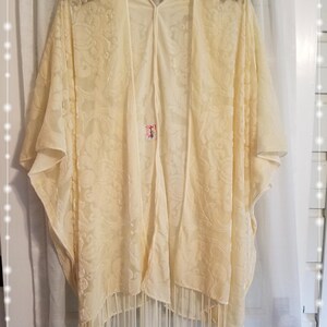 May include: A cream-colored, open-front kimono with a floral pattern and fringe trim. The lightweight fabric drapes loosely, and the garment hangs on a decorative gold hanger against a white curtain backdrop. The kimono is a stylish layering piece.