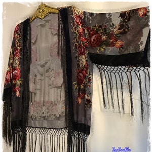 May include: A sheer, gray kimono-style garment with a floral pattern of red roses and green leaves. The kimono has black velvet trim and long black fringe along the edges. It is hanging on a gold-colored hanger.
