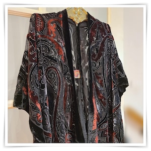 May include: A dark gray and burgundy paisley patterned kimono-style robe with black velvet trim. The robe is hanging on a gold-colored hanger. The paisley pattern is detailed with silver-colored embellishments.