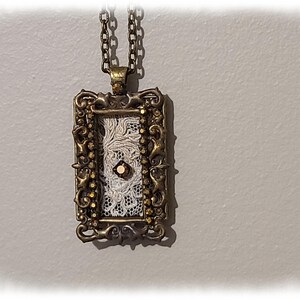 May include: A vintage-style pendant necklace with a rectangular frame. The frame is bronze-colored with ornate detailing and small, gold-colored accents. Inside the frame is a white lace design with a central, small, round, brown gem. The necklace chain is also bronze-colored.