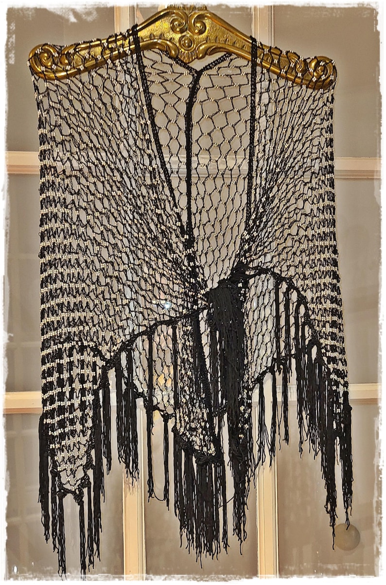 May include: A black, open-weave shawl with a decorative fringe. The shawl is adorned with small, off-white beads and hangs from a gold-colored, ornate hanger. The design features a net-like pattern.